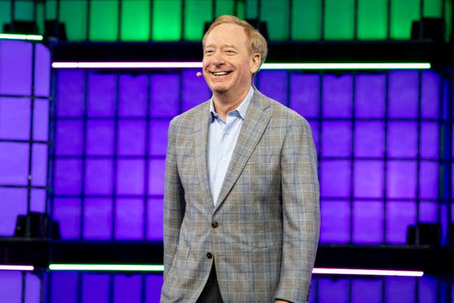 Brad Smith wearing a plaid grey blazer, laughing, while on stage in front of panels lit up in green and purple