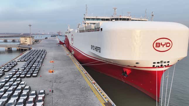 aerial view of rows of electric vehicles on a dock beside a large white and red ship in the water beside the port