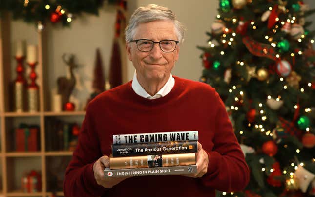Bill Gates holding four books, wearing a red sweater and white button down shirt in front of a Christmas tree